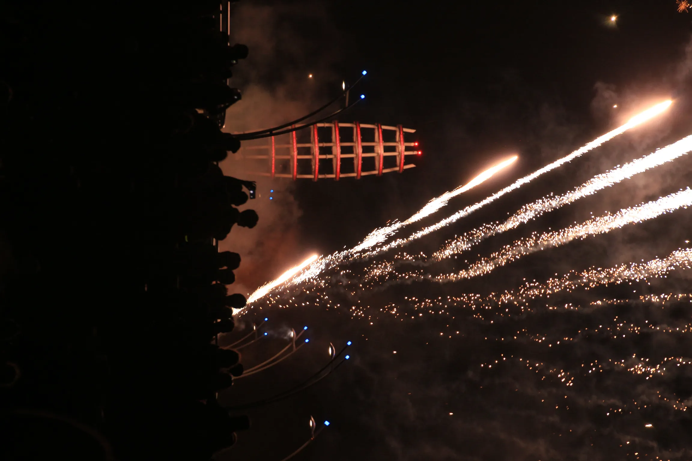 Fireworks streaming out over Lake Ontario at night beside the Brant Street Pier, its lattice tower lit red, with a crowd silhouetted along the shore.