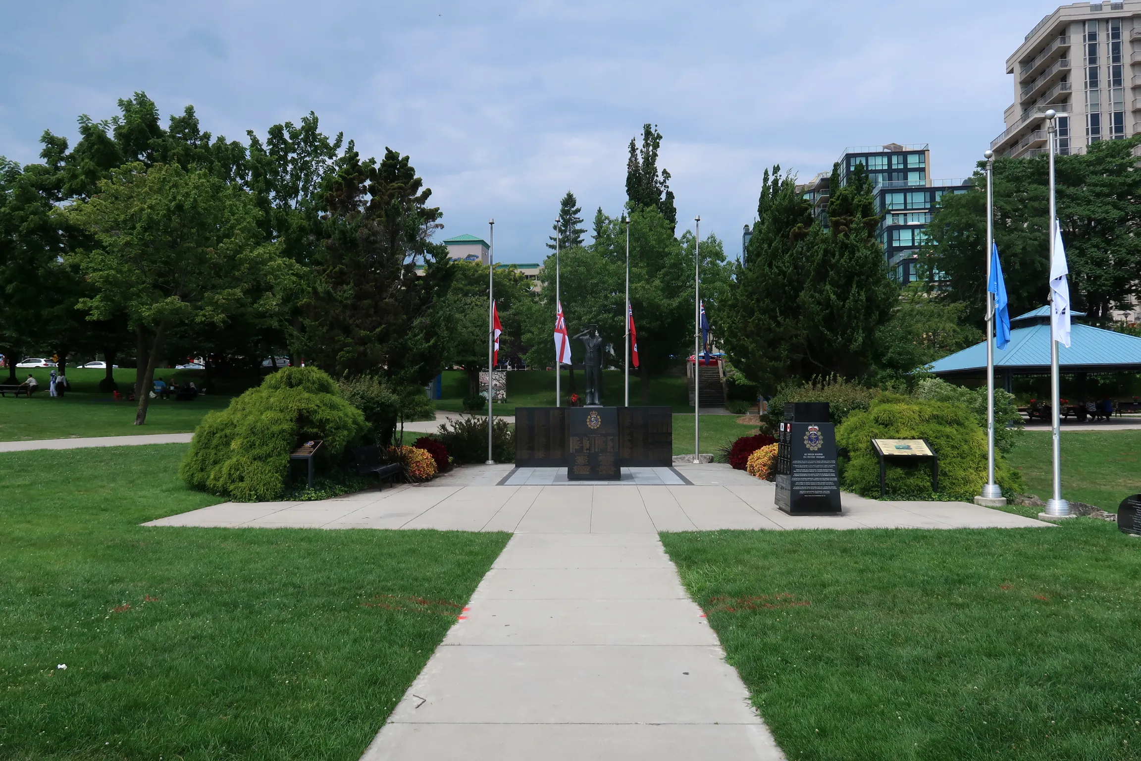 The dark granite Naval Ships' Memorial Monument standing between flagpoles, flanked by autumn chrysanthemums and evergreens, with a walkway leading to it.