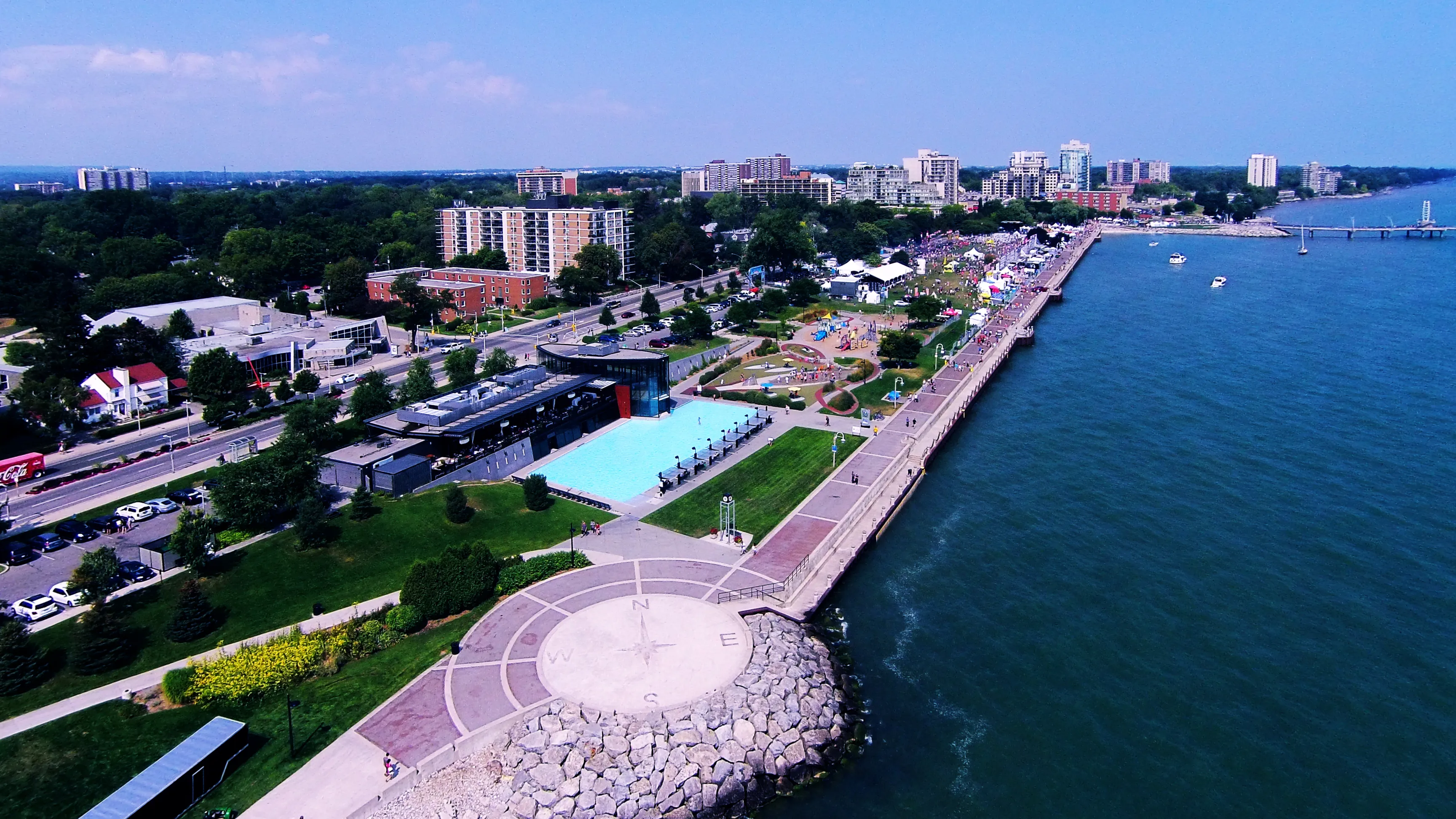An aerial view along the waterfront: the brick promenade curving beside the lake, the compass plaza and a blue-roofed pavilion below, apartment towers and the pier beyond.