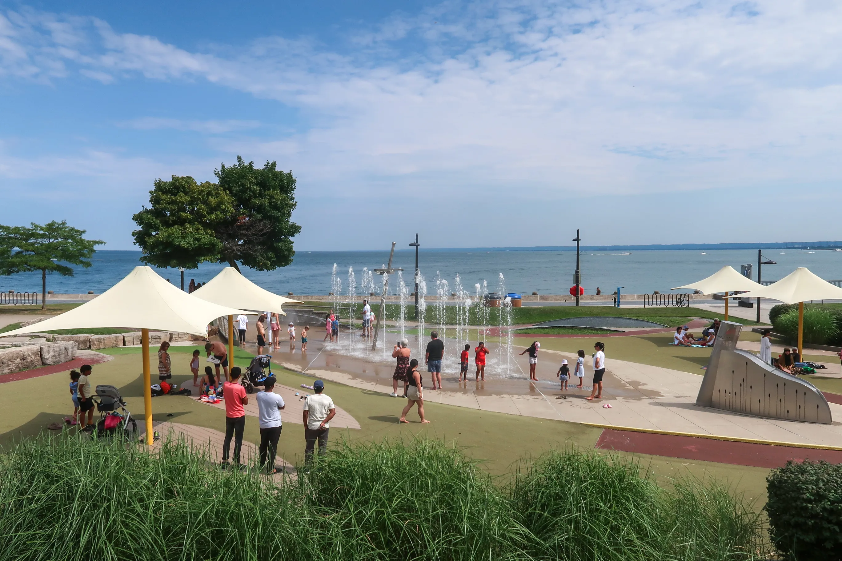 Children and adults spread across the splash pad as vertical jets of water rise from the pavement, white sail shades overhead and Lake Ontario behind.